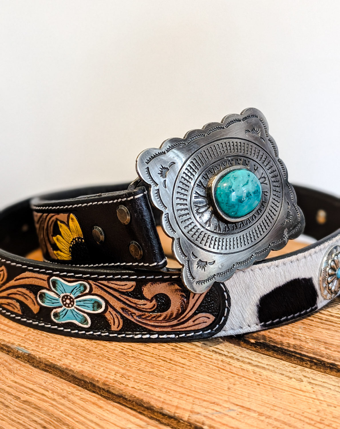 A floral tooled western belt with a prominent silver buckle featuring a turquoise stone, and sections with embroidered floral patterns in blue and yellow on a black background, alongside a section with a black and white cowhide texture.