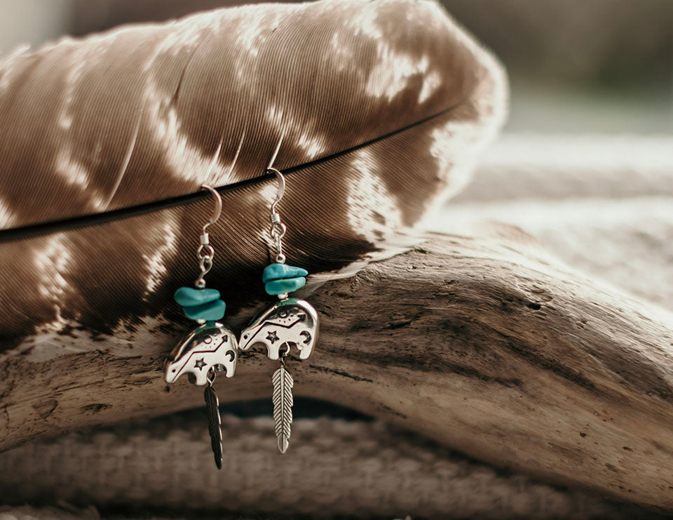 A super cute bear design hanging earrings with chunks of genuine turquoise nuggets and feather detailing for those that love a piece with spiritual significance. The delicate bear pendant is engraved with tiny tribal motifs. Earrings showcased on a natural brown feature background. 