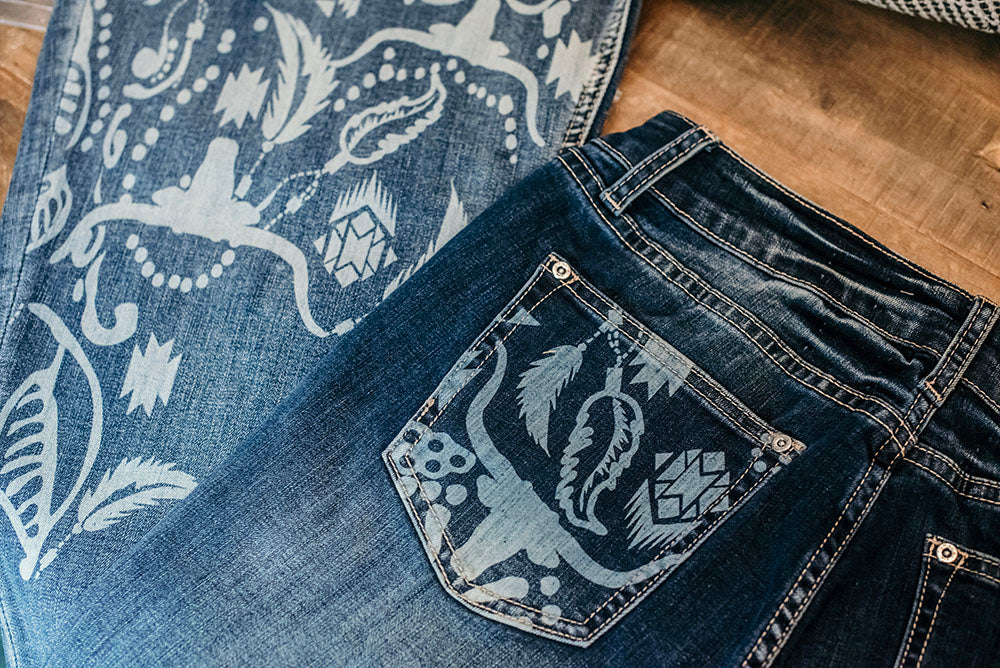 The back pockets and bottom of the leg of a medium blue pair jeans with a bleached out buffalo and aztec print. 