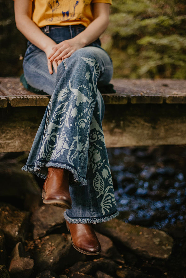 Woman wearing A super fun and sexy jeans design with a bleached out buffalo and aztec print on the back pockets and lower leg. She is shown sitting on a wooden bridge. UK Exclusive Grace in LA Stockist, Get Grace in LA in UK. 