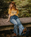 Woman wearing a beautiful yellow top paired with A super fun and sexy jeans design with a bleached out buffalo and aztec print on the back pockets and lower leg. She is shown sitting on a wooden bridge.