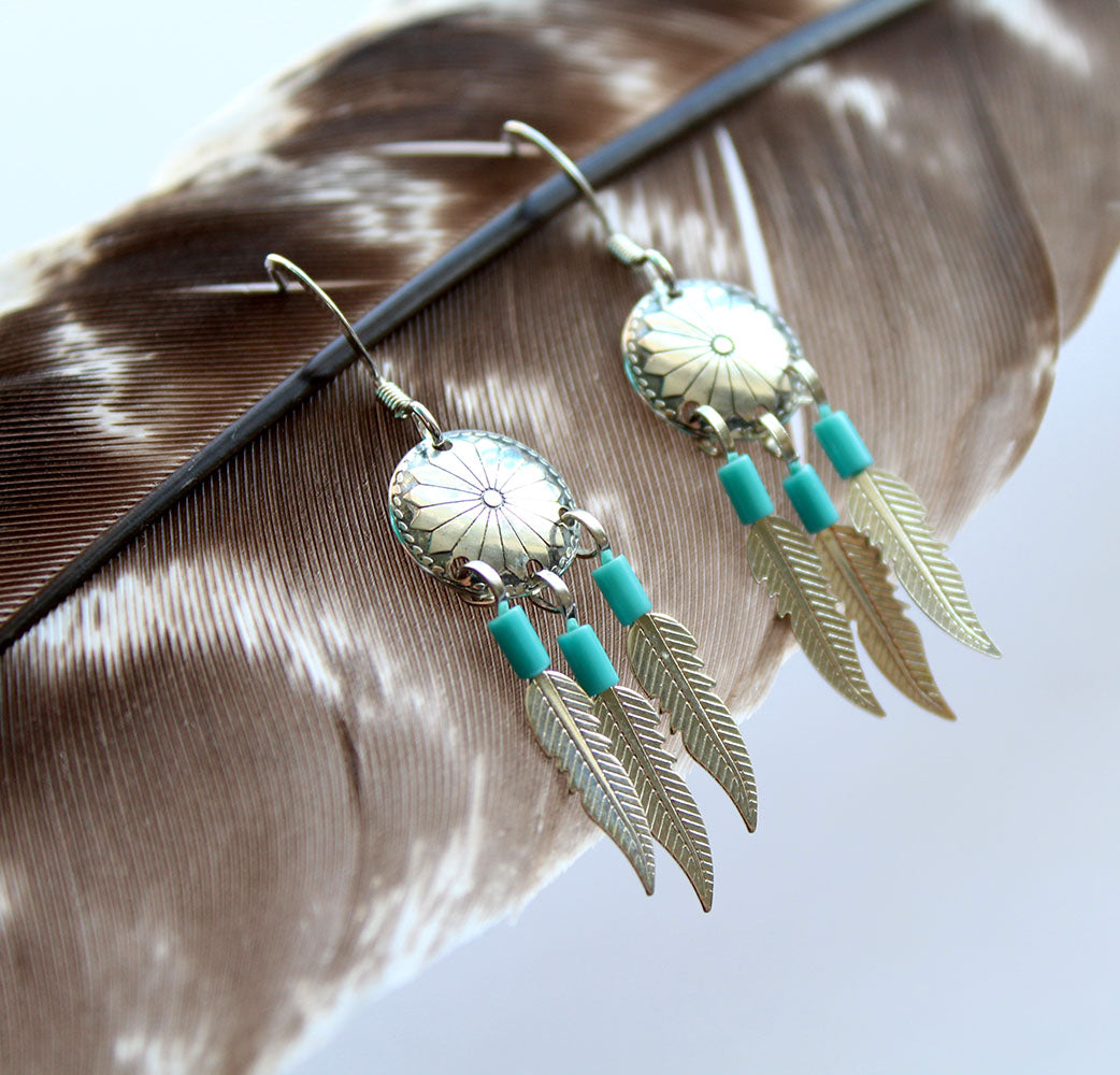 A classic Native American design. These earrings feature tripple dangling silver feathers  topped with Turquoise beads hanging from an intricate concho charm. The earrings are a hook style and are handmade by a Native American Artist. They are showcased on a brown feather background.